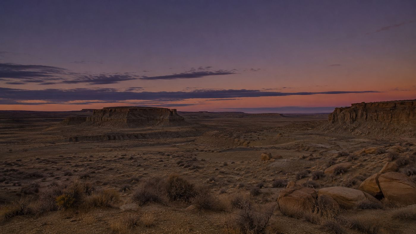 The wide arid desert canyon landscape of Chaco Canyon at dusk, low mesas and dry scrub under a vast darkening sky