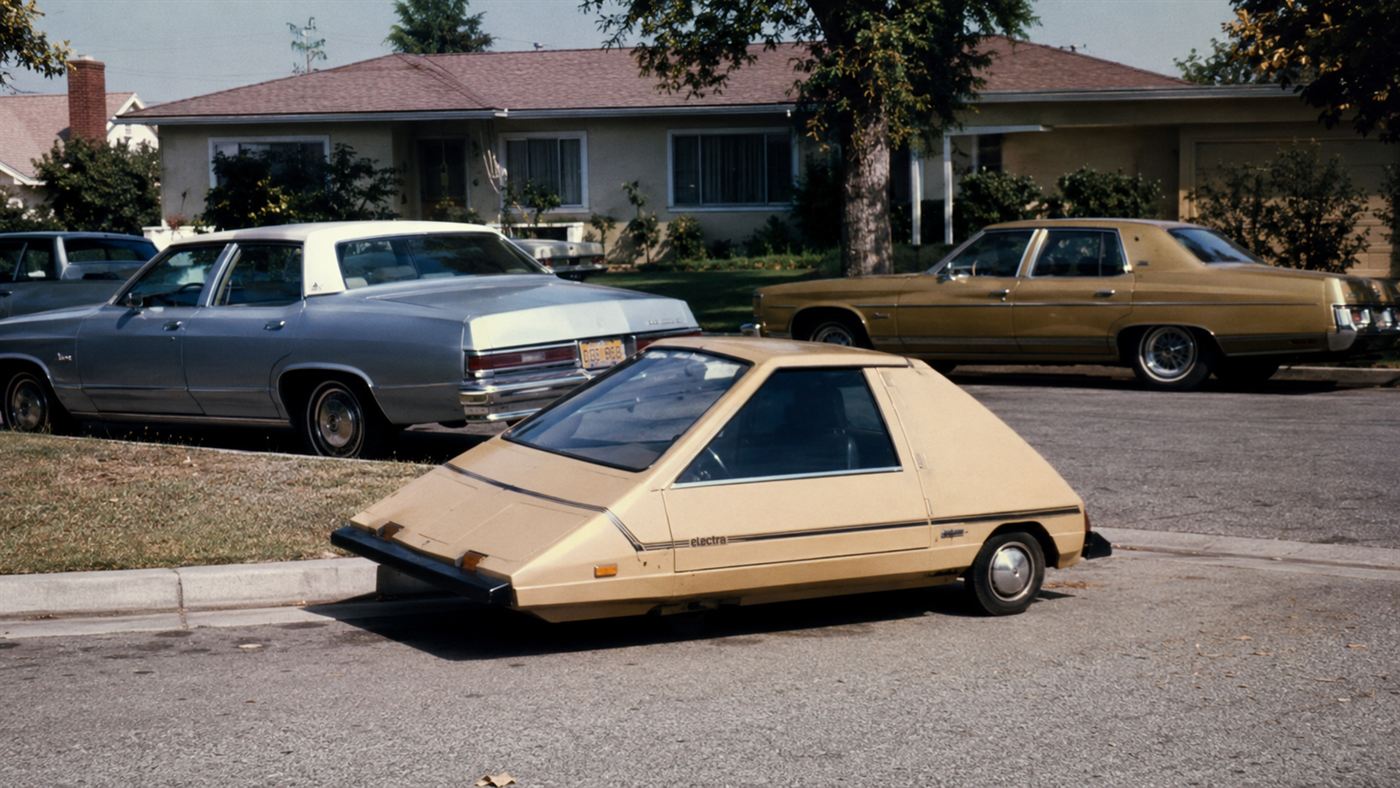 A 1974 CitiCar, a tiny wedge-shaped plastic electric car, parked on an American suburban street