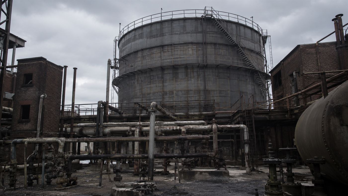 A large cylindrical 1940s industrial gas storage tank in an urban plant yard under an overcast sky