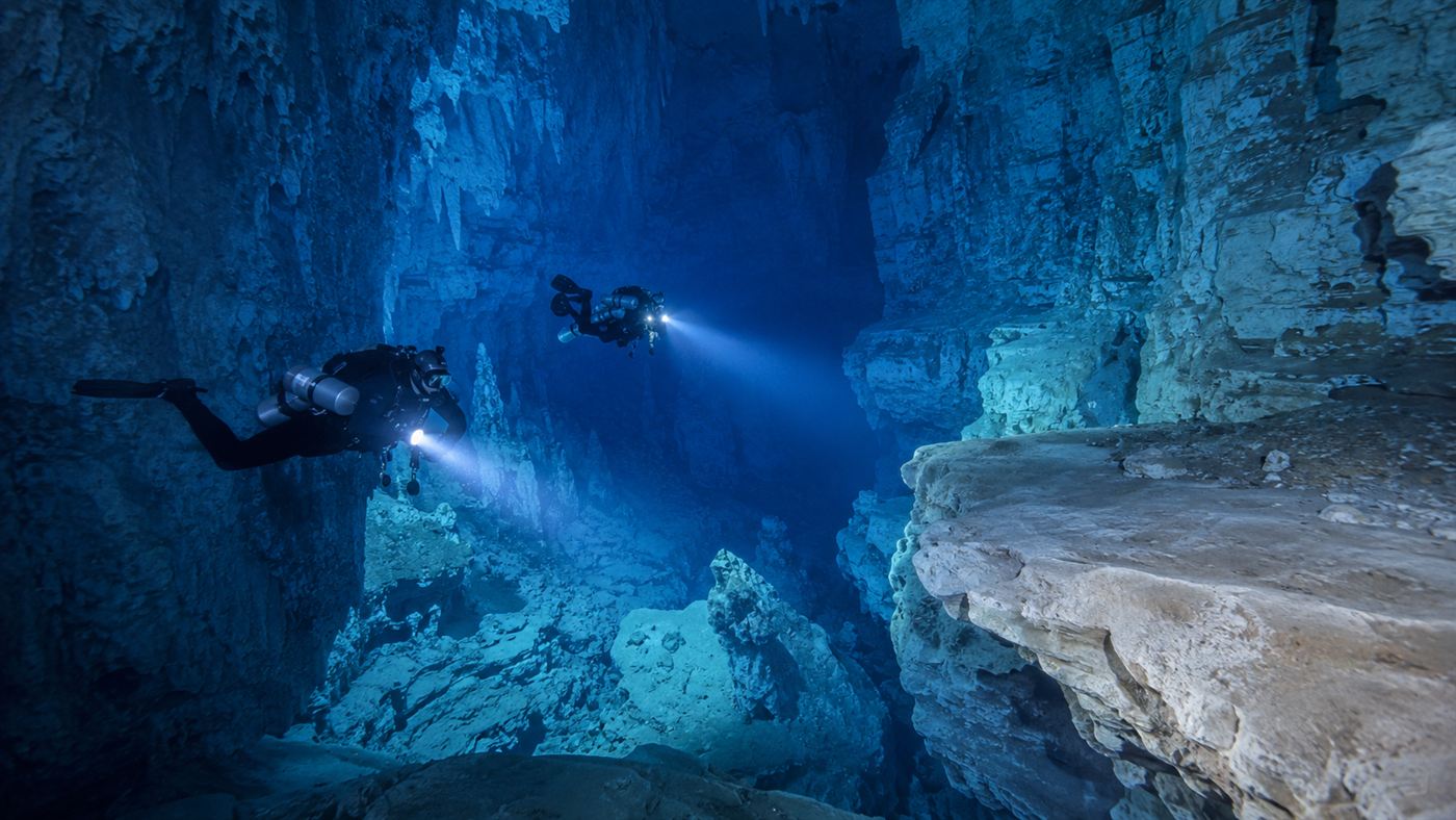 Two scuba divers with lights descending into the deep clear blue flooded limestone cavern of Devils Hole to count fish