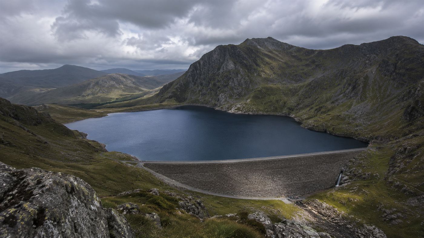 A high mountaintop reservoir of deep blue water held by a dam, surrounded by rugged grey Welsh mountains under a moody sky