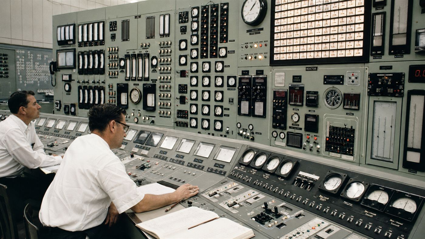 A 1950s nuclear reactor control room with rows of analog dials and switches and scientists at the controls