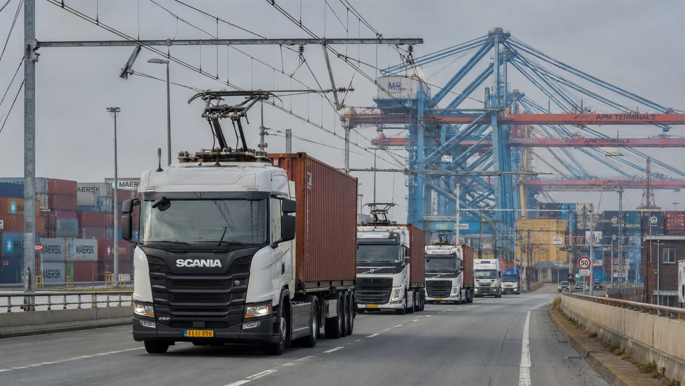 A line of trucks under overhead electric wires on a road near a busy container port with cranes in the background