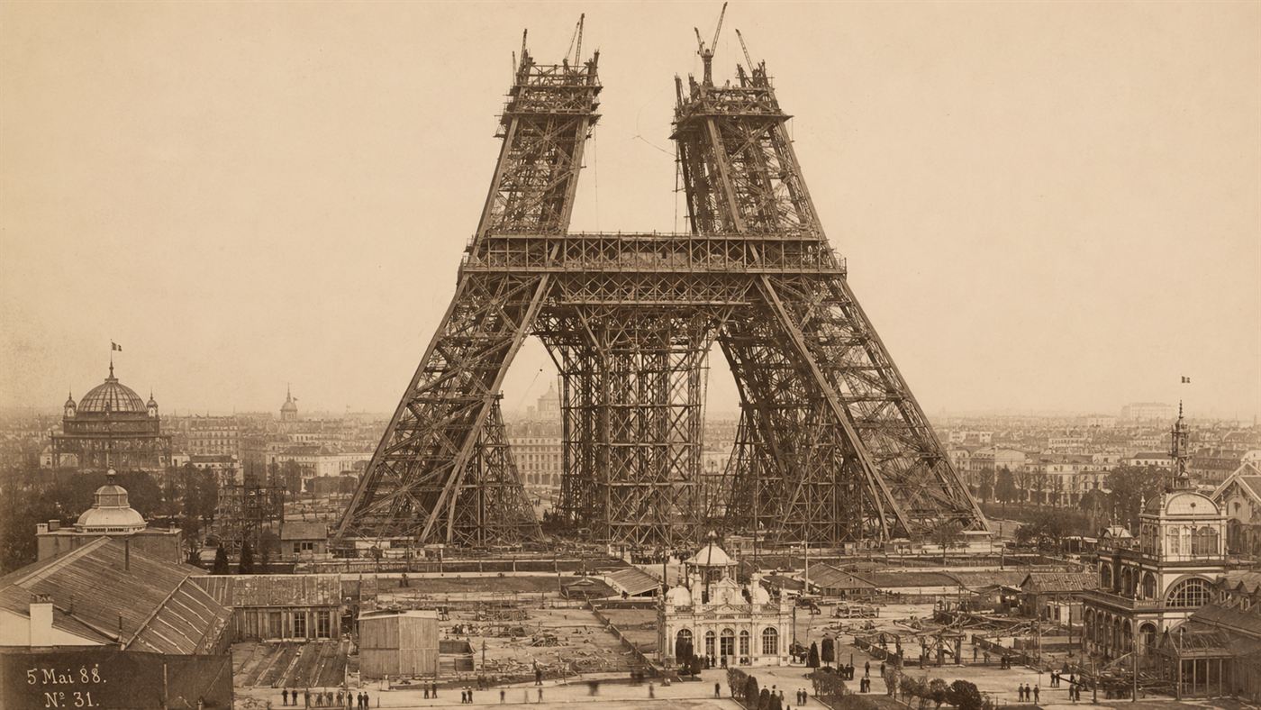 The half-built Eiffel Tower in 1888, its iron lattice legs rising over the Paris exhibition grounds in a sepia-toned scene