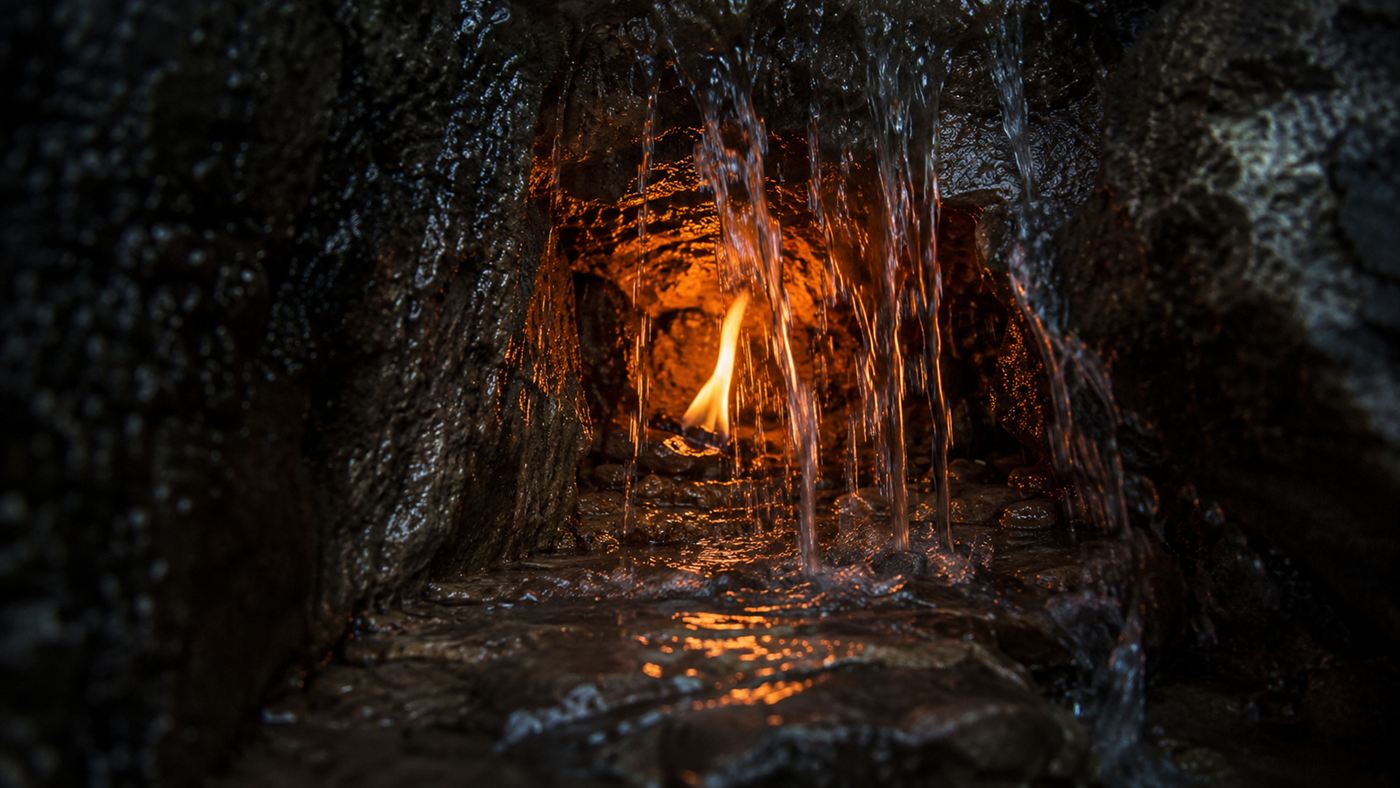 Close-up of a small orange flame flickering inside a dark wet rock alcove behind a veil of falling water