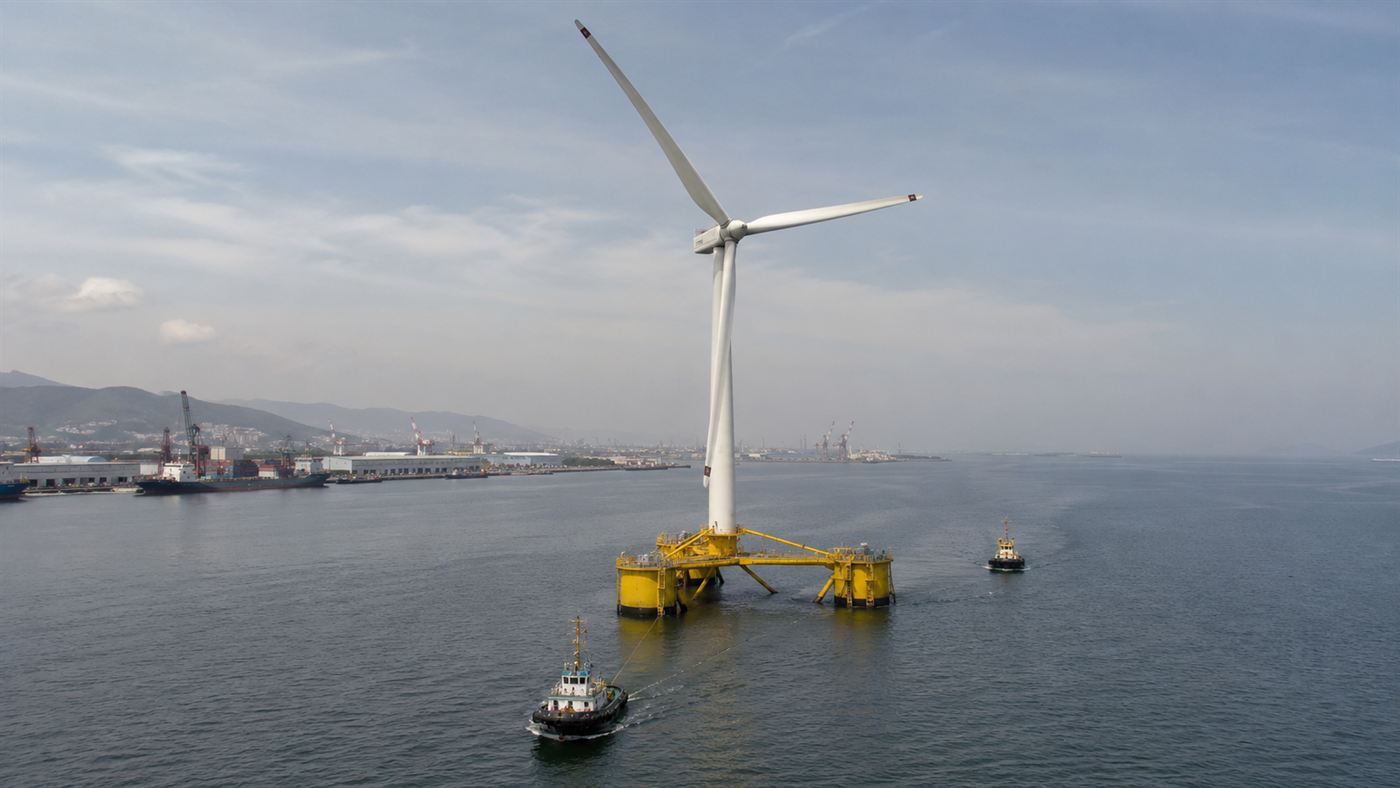A floating wind turbine on its spar being towed out to sea by tugboats from a harbour