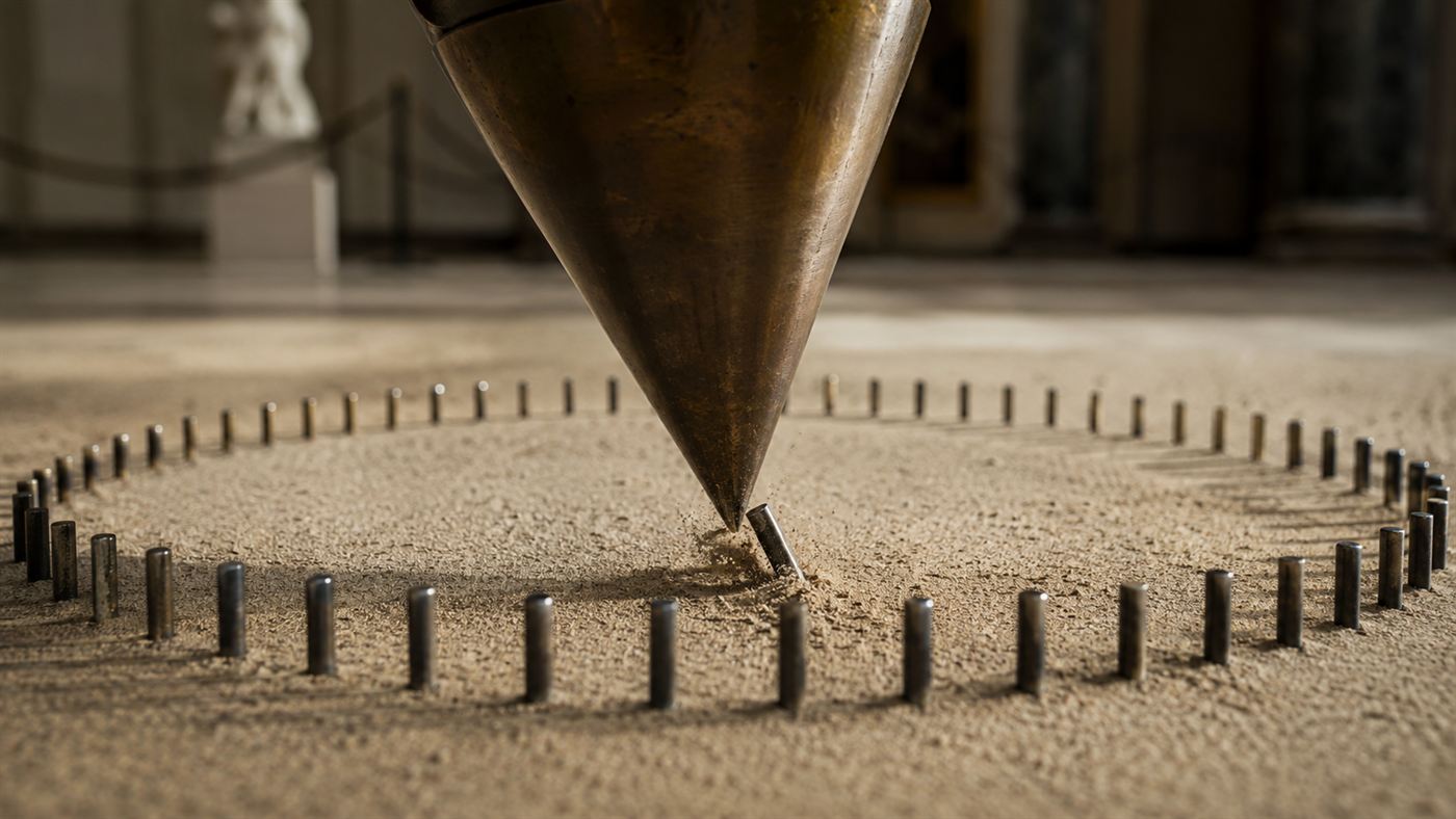 The pointed tip of a pendulum bob sweeping over a ring of small pins on a sand floor, knocking one over