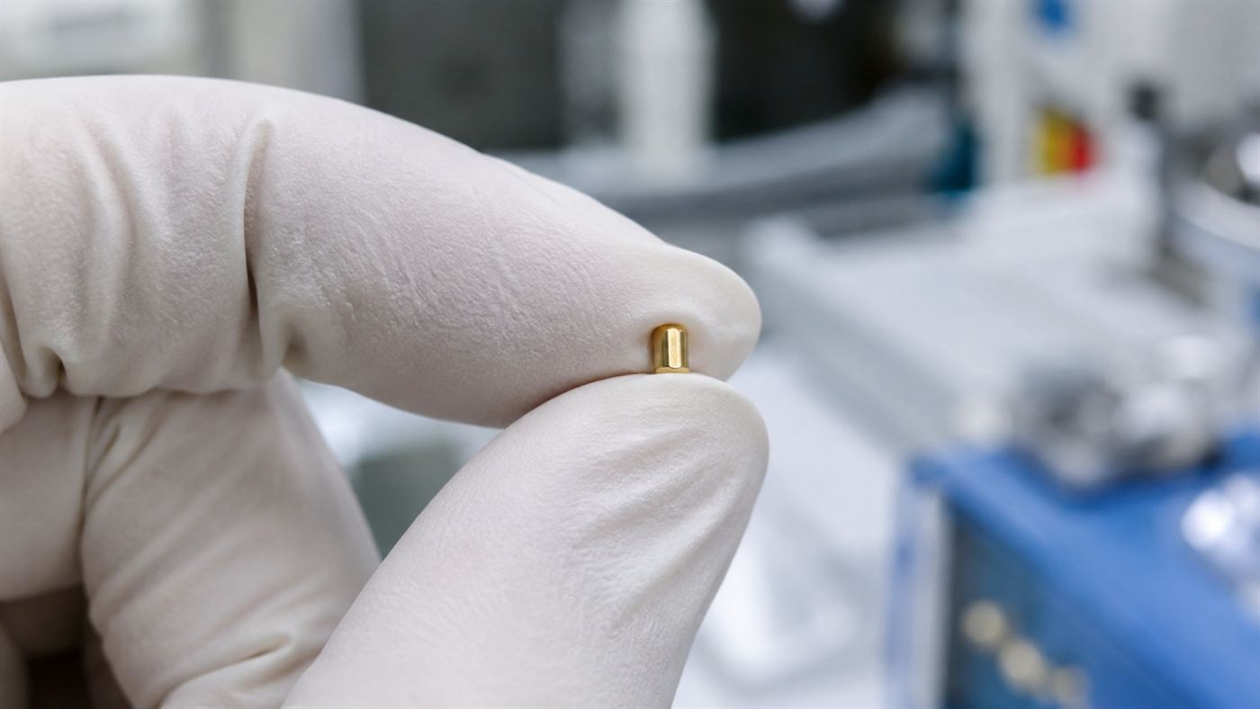 A gloved hand holding a tiny peppercorn-sized gold fuel capsule used in laser fusion experiments