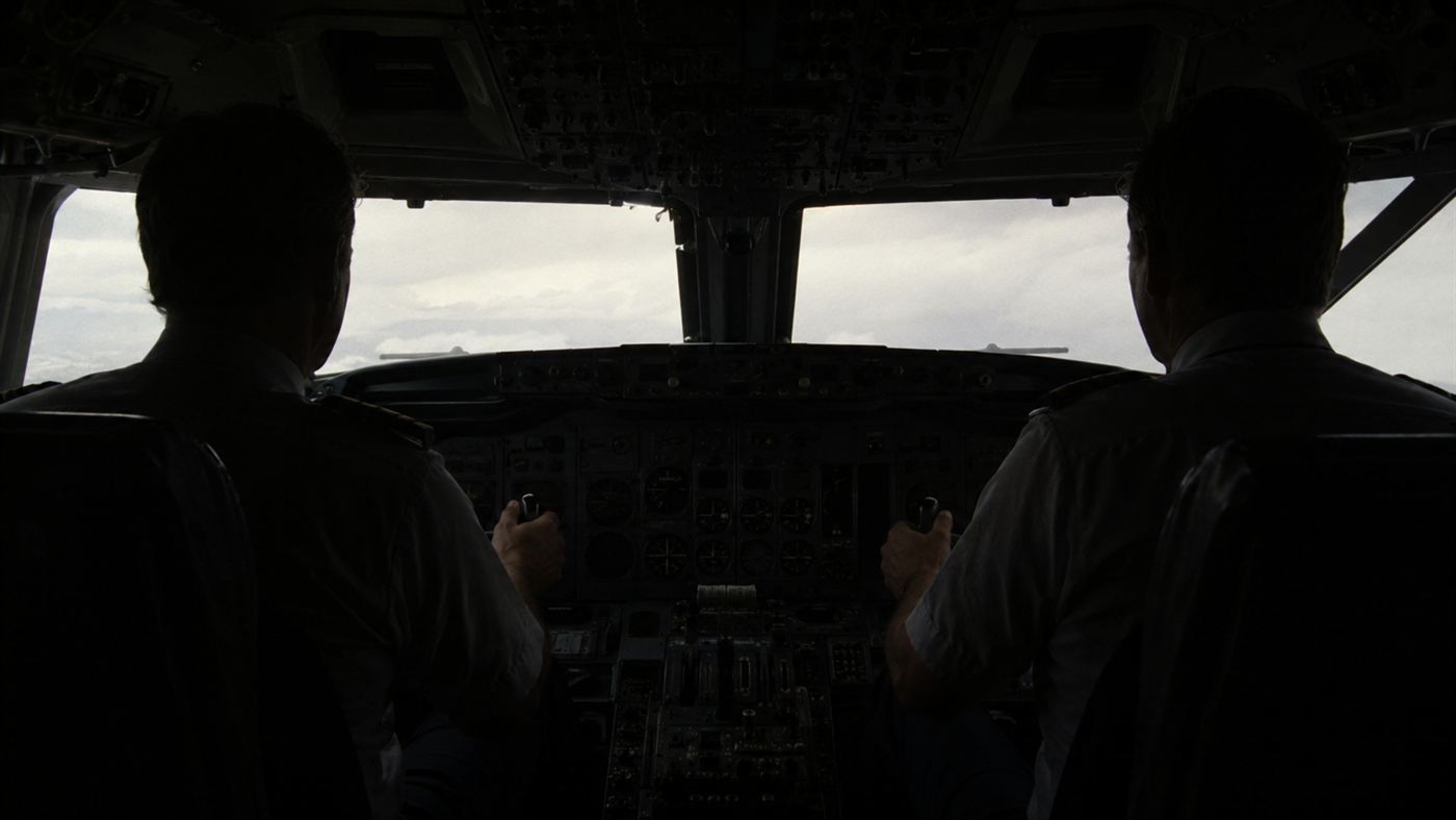 A dim 1980s airliner cockpit with dark, unlit instrument panels and two pilots in silhouette