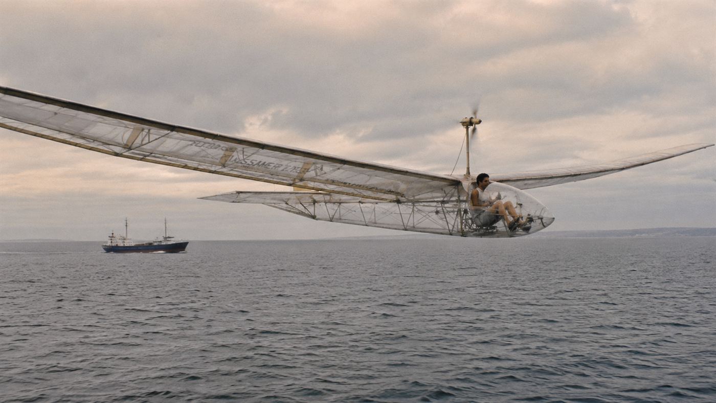 A human-powered aircraft with very long thin wings flying low over open grey sea, a pilot pedalling inside