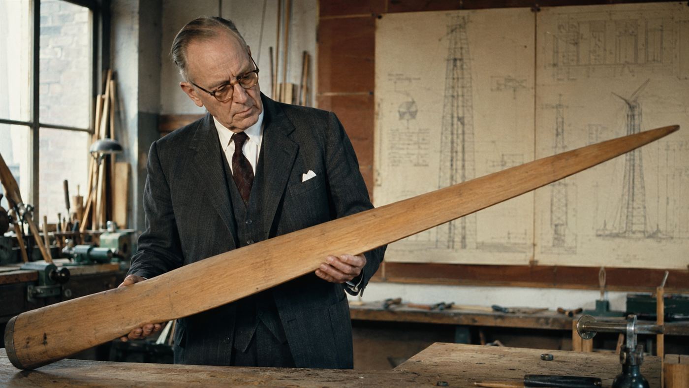 An elderly Danish engineer examining a long wind-turbine blade in a 1950s workshop with blueprints