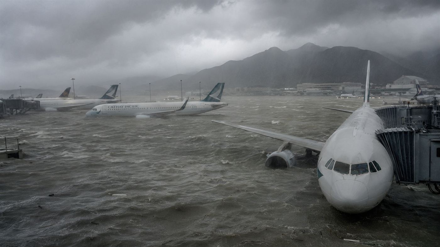 An island airport runway flooded by grey storm seawater during a typhoon, parked airliners standing in floodwater up to their engines under a dark stormy sky