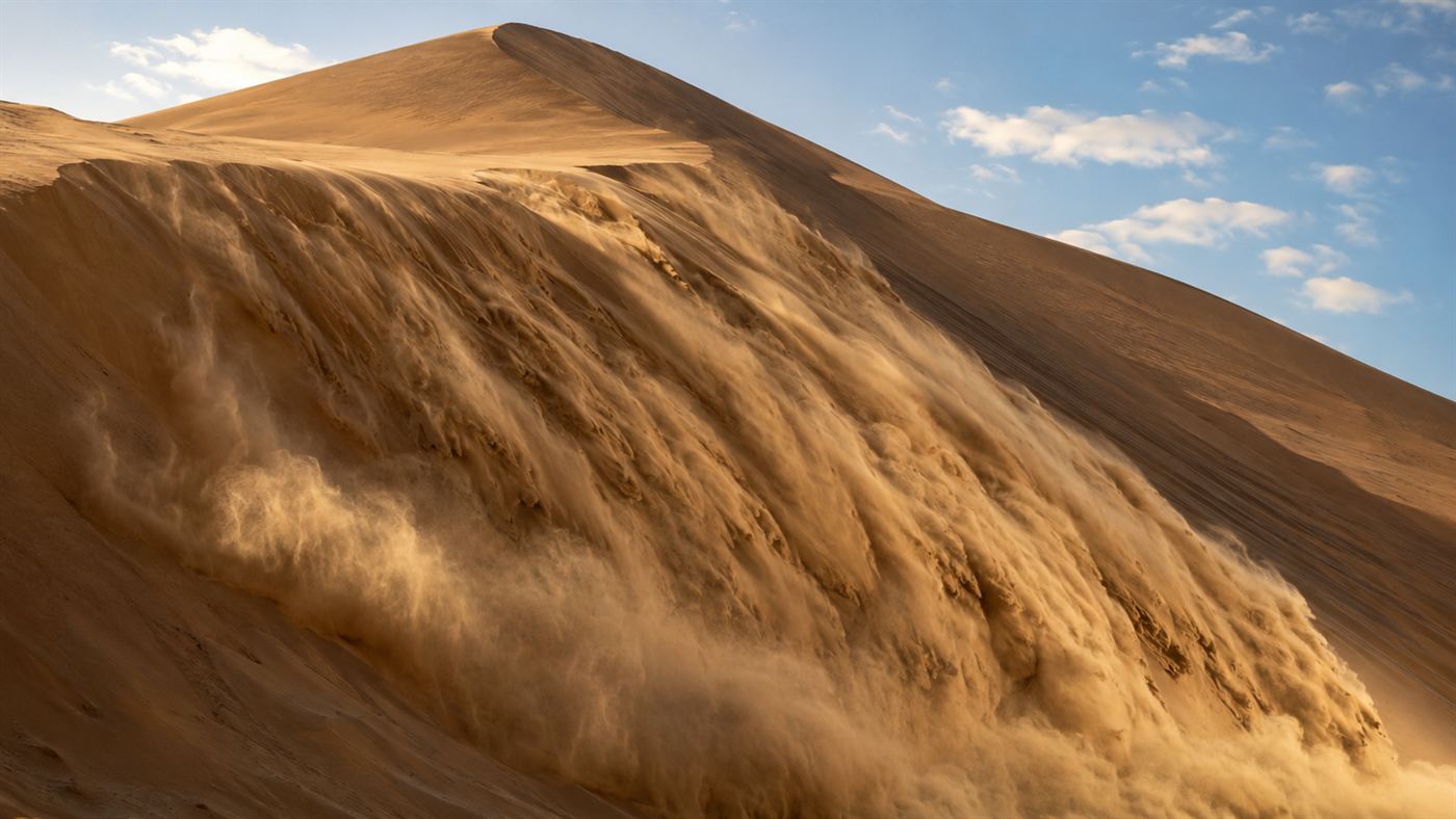 A sheet of dry sand avalanching down the steep face of a tall desert dune, sending up a faint haze