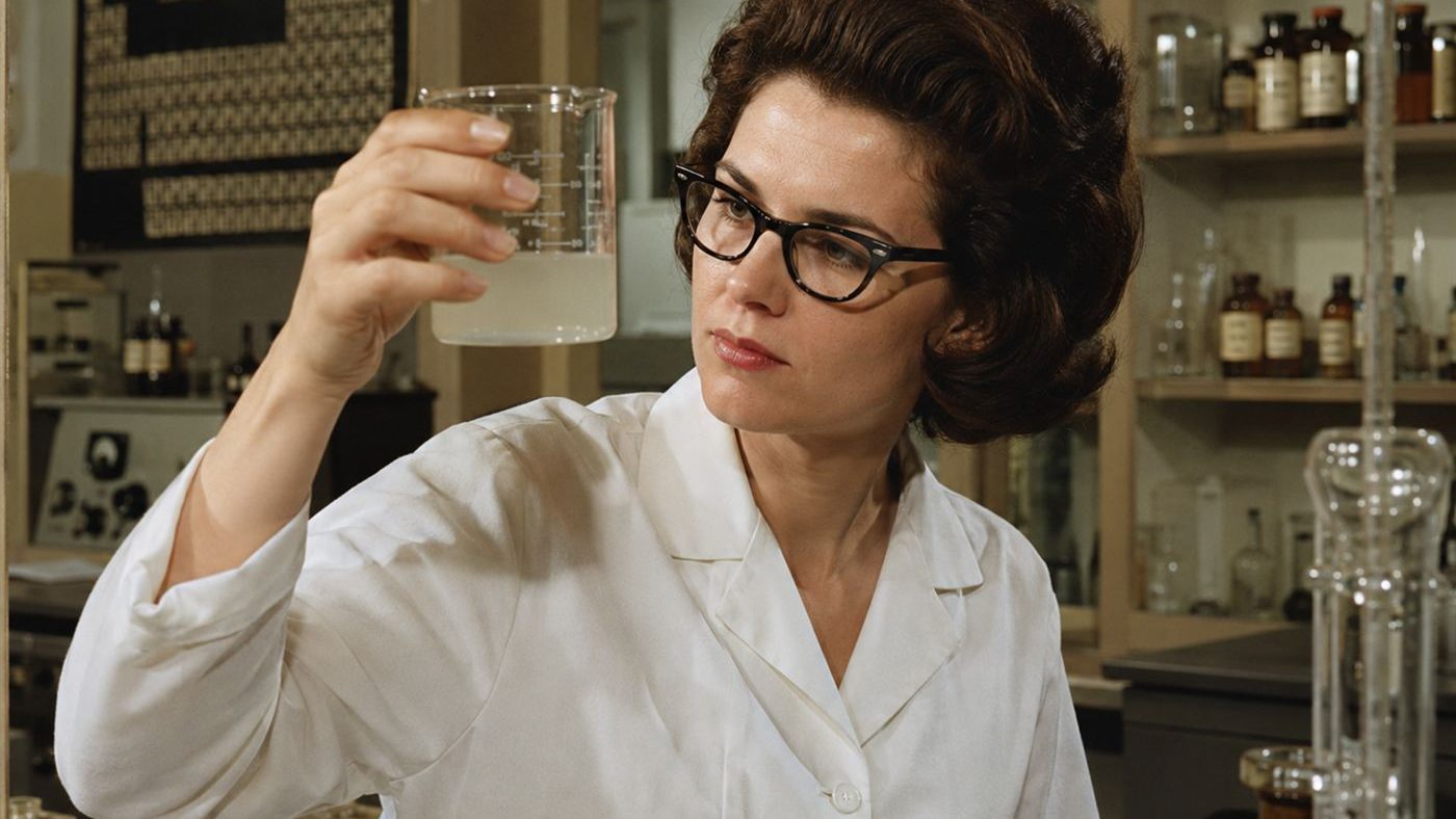 A 1960s woman chemist holding up a beaker of cloudy liquid, like Stephanie Kwolek discovering Kevlar