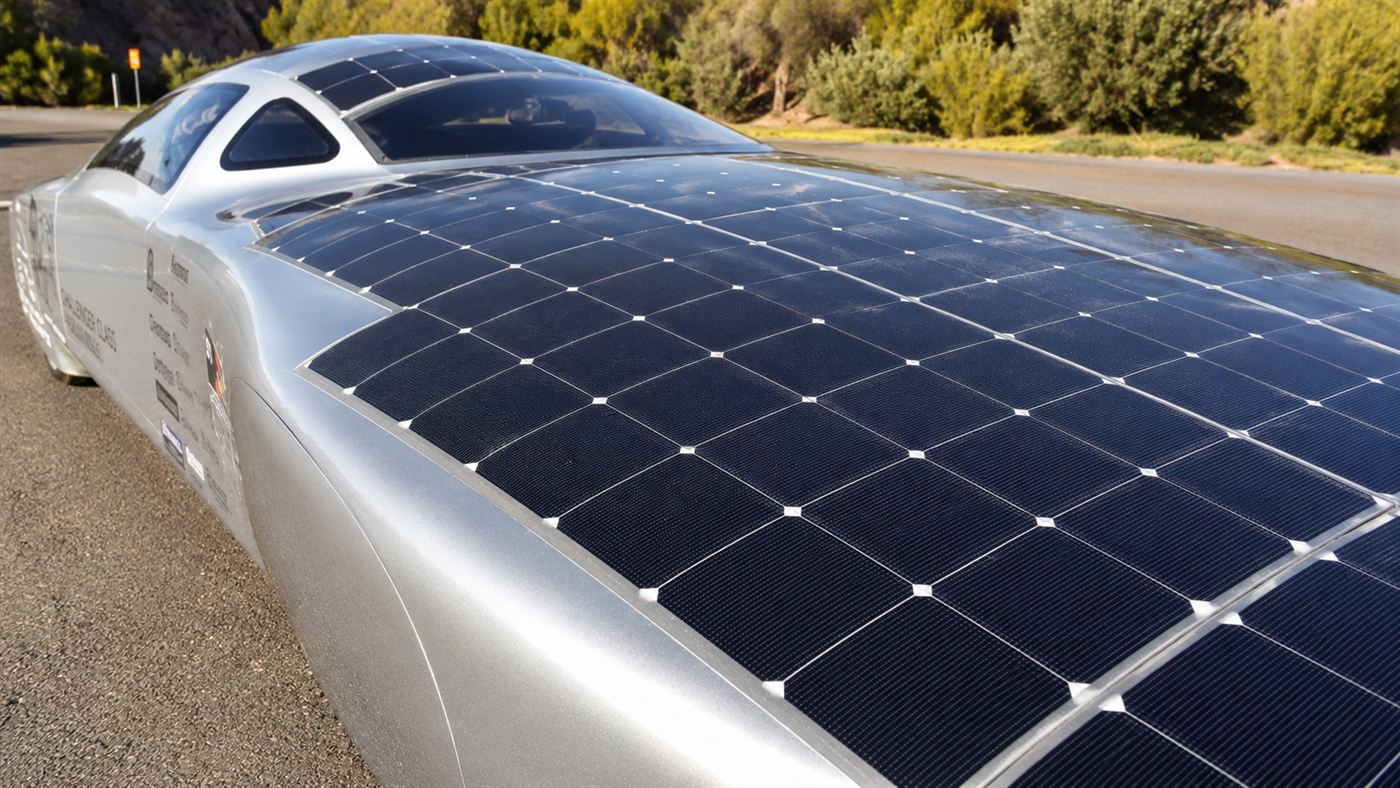 Close view of the long curved roof and bonnet of a sleek silver car densely covered in hundreds of dark photovoltaic solar cells reflecting the sky