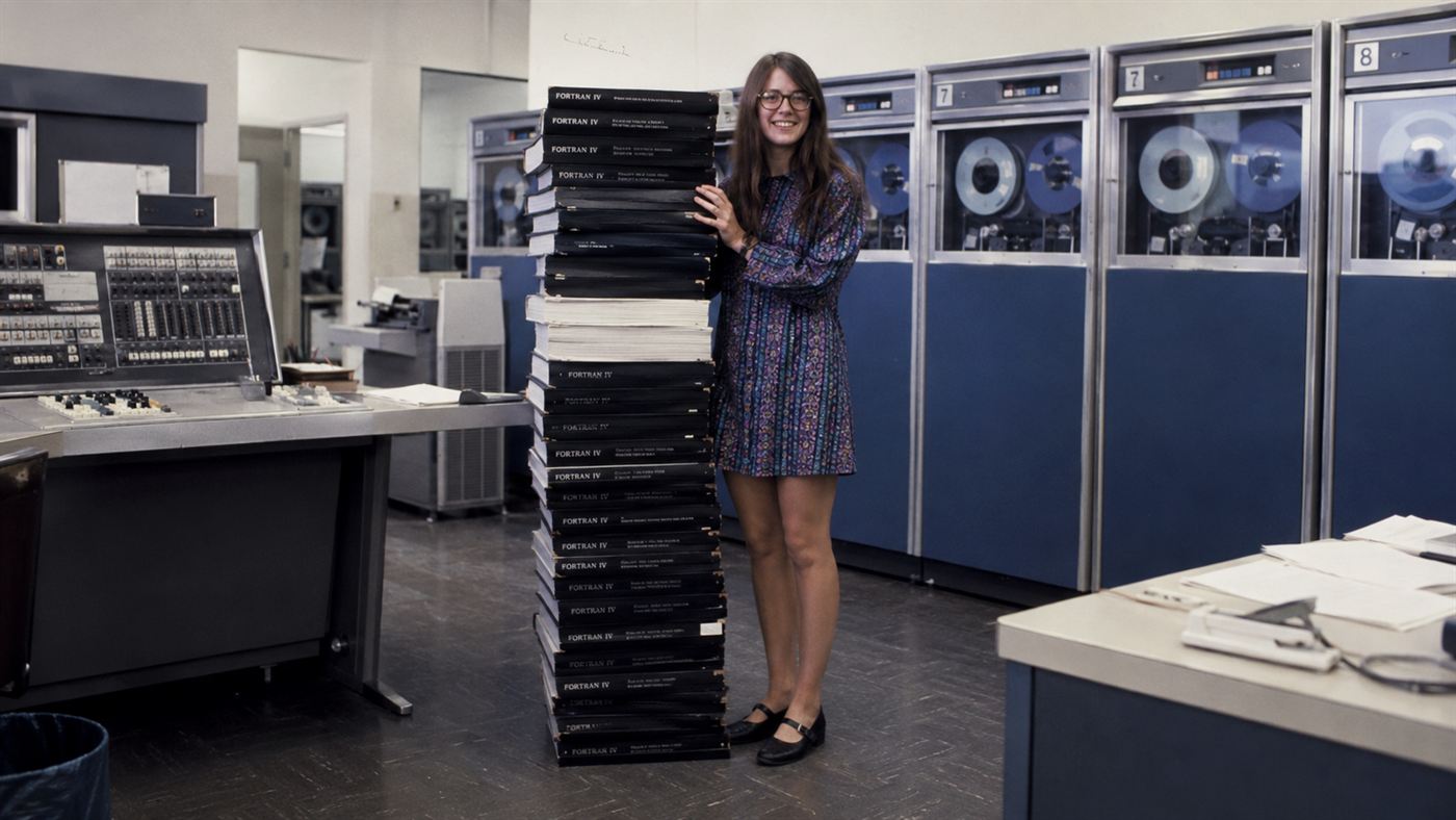 A 1960s woman scientist beside a tall stack of printed Apollo source code, evoking Margaret Hamilton
