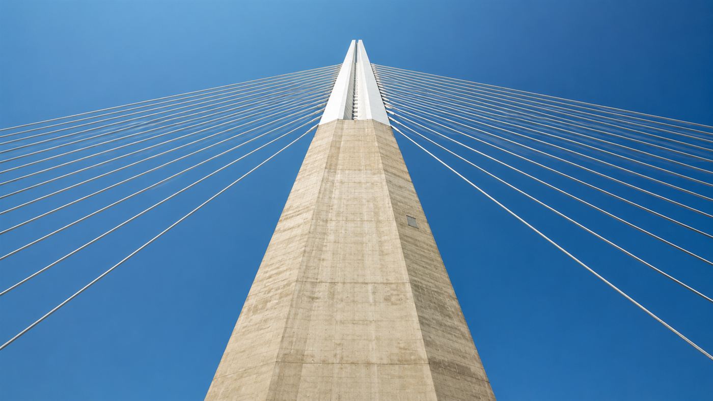 Looking up at one of the tall tapering concrete piers and white mast of the Millau Viaduct against the sky