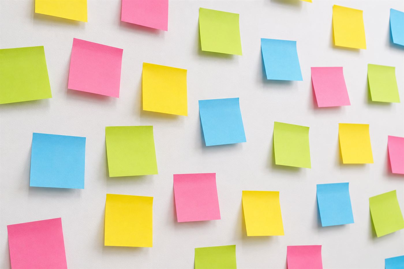 A cluster of bright yellow, pink and blue Post-it notes stuck at angles on a plain wall, some curling at the corners