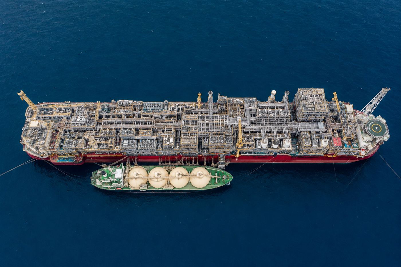 An aerial view looking down the full length of the Prelude FLNG vessel, its deck packed with processing units and pipework, dwarfing a carrier ship alongside