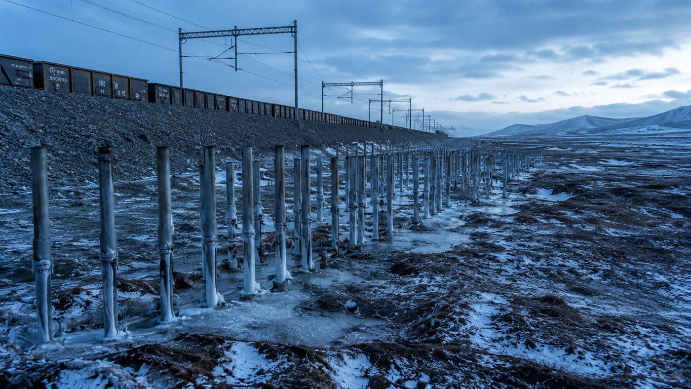 Rows of metal cooling pipes standing in the ground beside a railway embankment on a frozen plateau to keep the permafrost cold