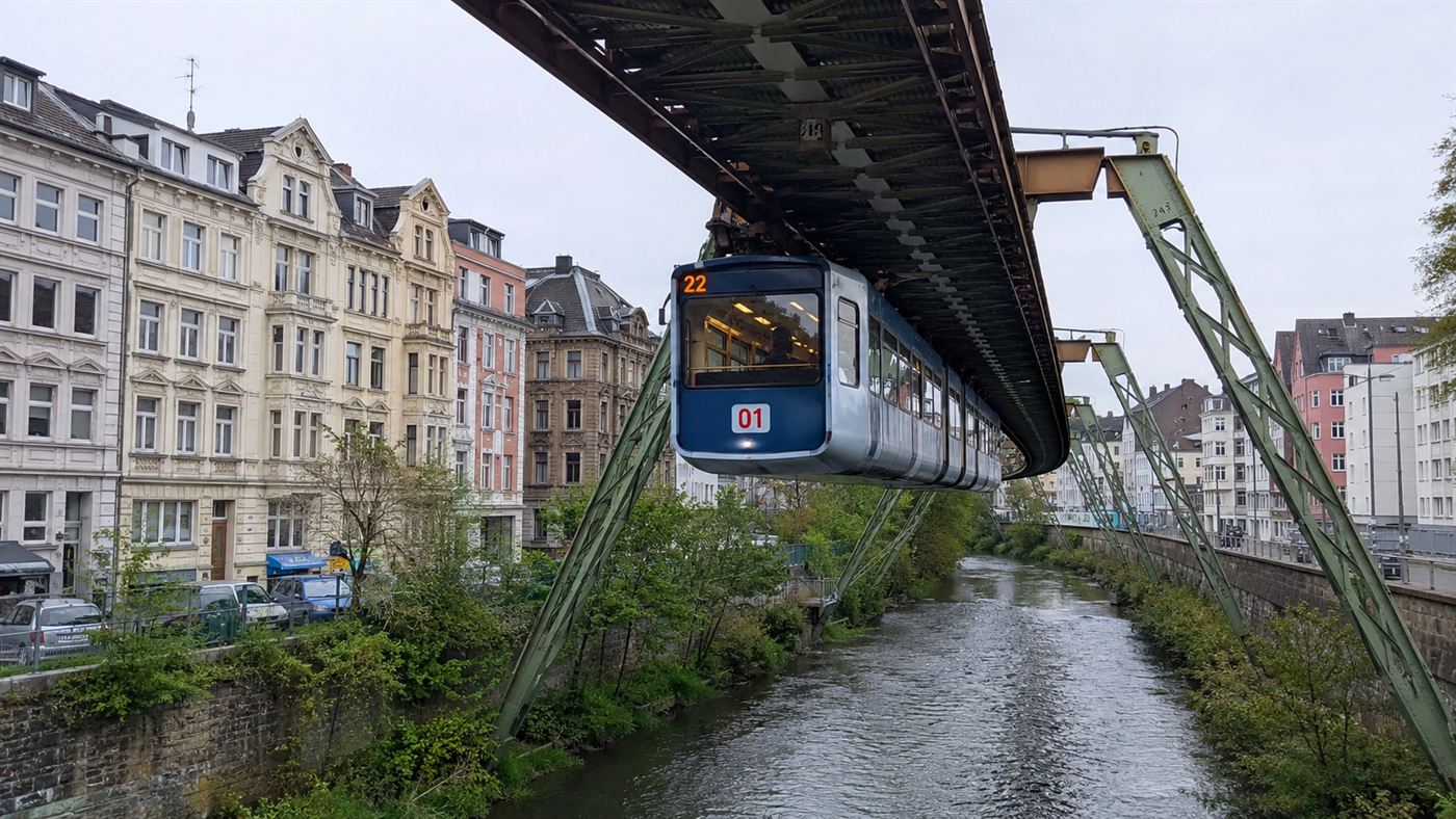 A blue and silver Wuppertal Schwebebahn carriage hanging from a steel frame above the Wupper river with old German buildings alongside