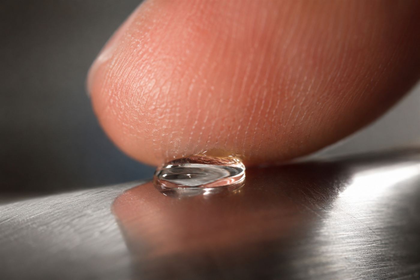 Extreme close-up of a single clear drop of Super Glue instantly bonding a fingertip to a metal surface under bright studio light
