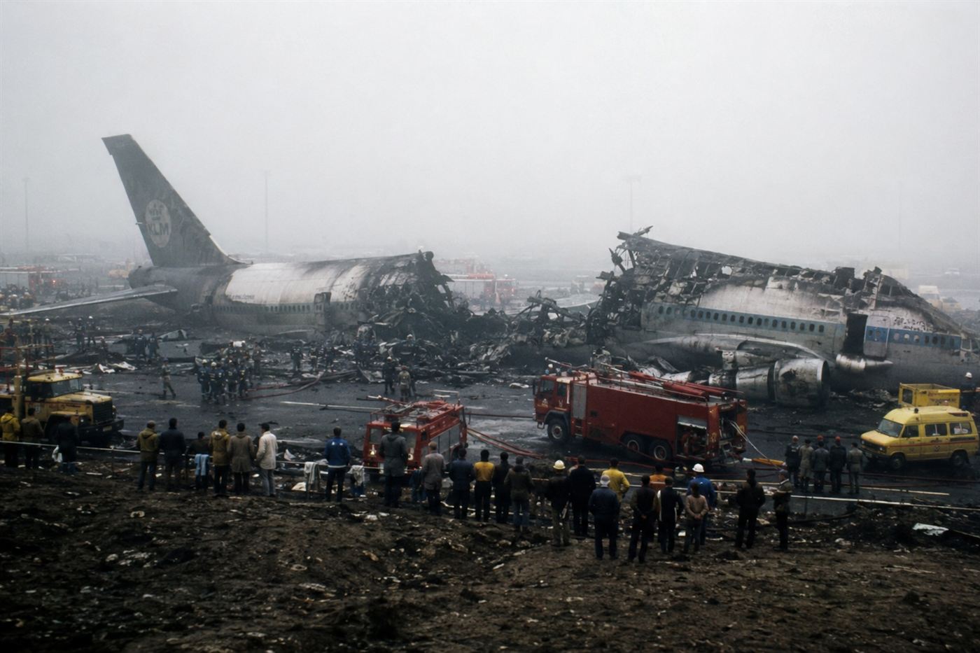 The aftermath of the Tenerife airport disaster in March 1977 showing the burned wreckage of the KLM and Pan Am Boeing 747s on the Los Rodeos runway, emergency vehicles surrounding the collision site, thick fog still visible over the airfield