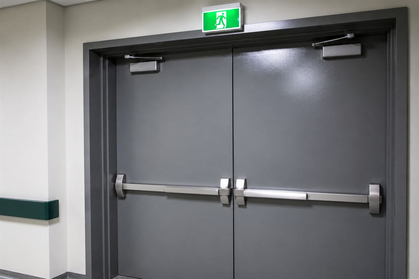 A modern theatre exit door fitted with a horizontal push-to-open panic bar and a lit exit sign above, a legacy of fire reforms