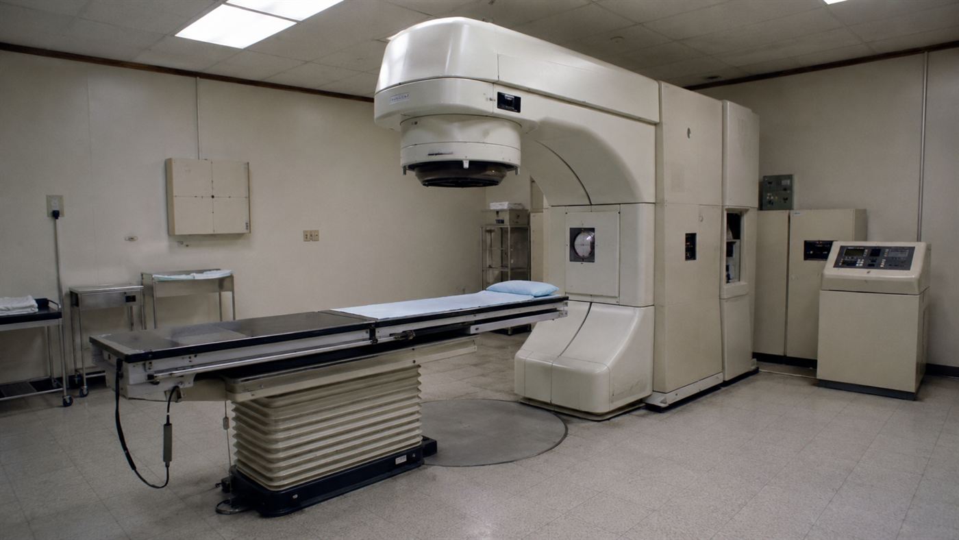 A 1980s Therac-25 style radiation therapy machine aimed at an empty treatment table in a hospital room