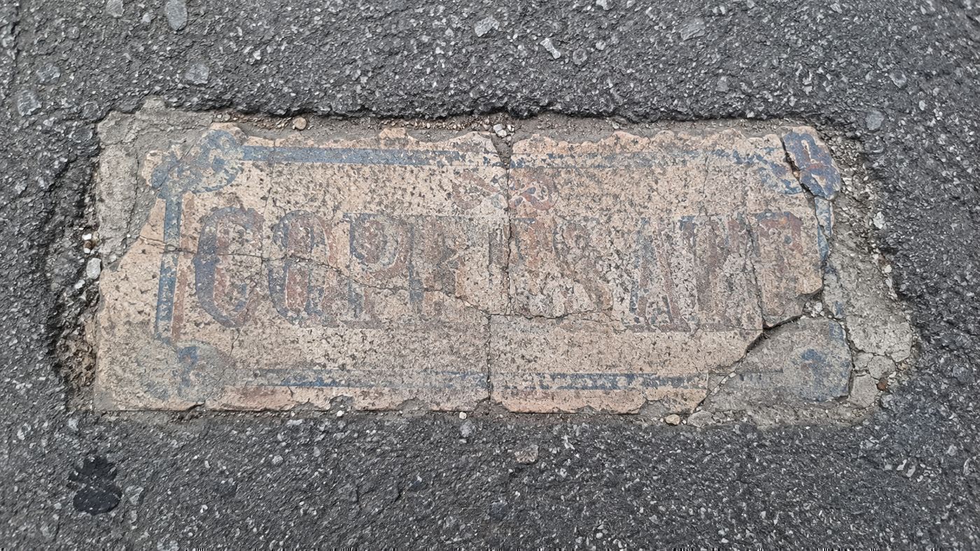 A worn handmade tile with faded lettering embedded flush in the cracked asphalt of a city street, a Toynbee tile