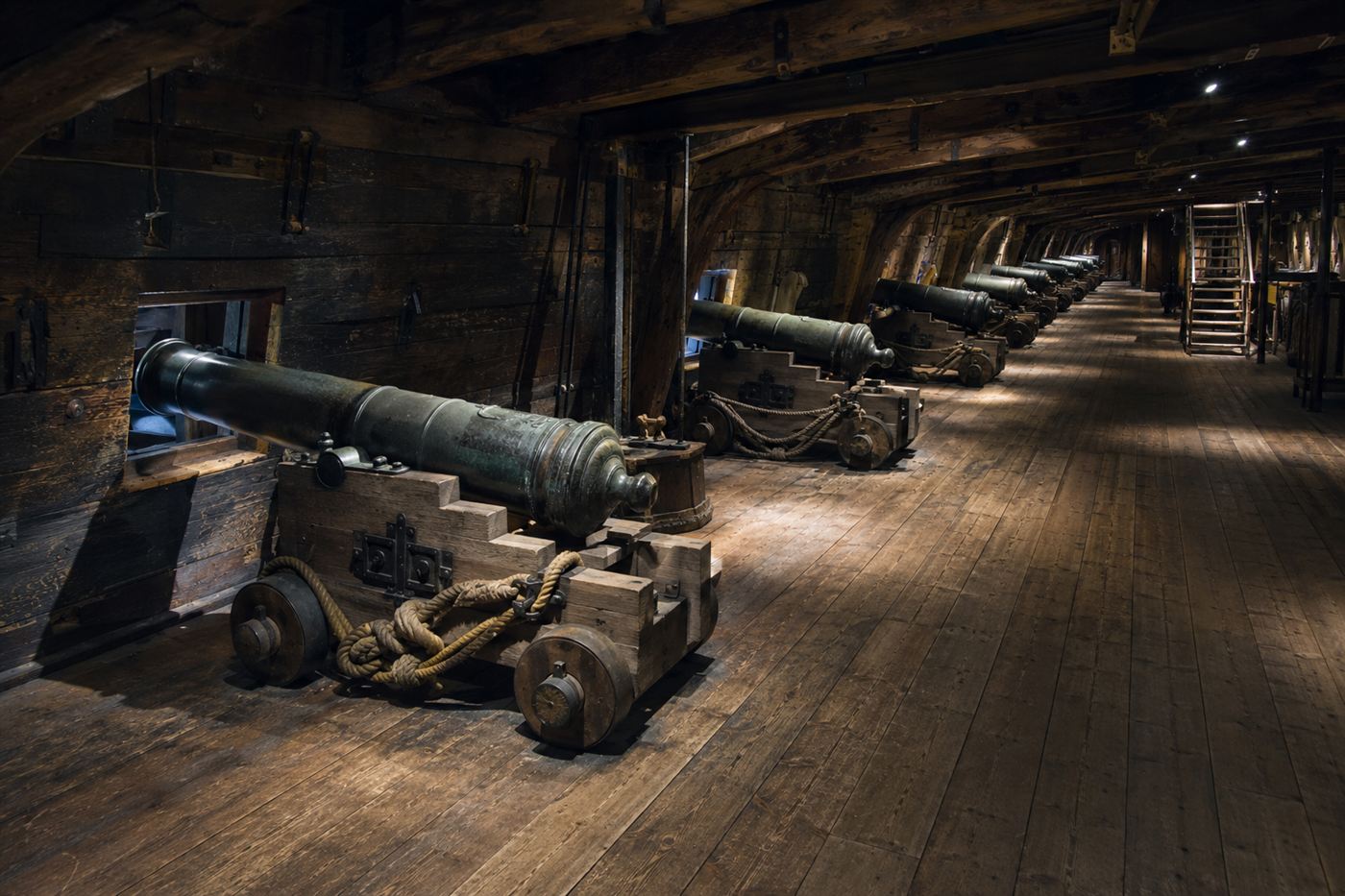 The preserved gun deck of the Vasa warship inside the Vasa Museum, rows of bronze cannons in their original positions along the lower gun deck, dim museum lighting showing the dark oak timbers