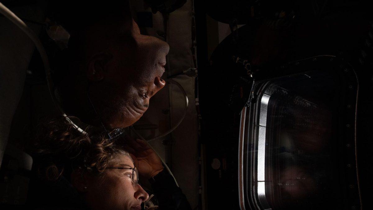 Christina Koch e Victor Glover, de perfil e na penumbra, olham juntos por uma janela da cápsula Orion durante o sobrevoo da Lua