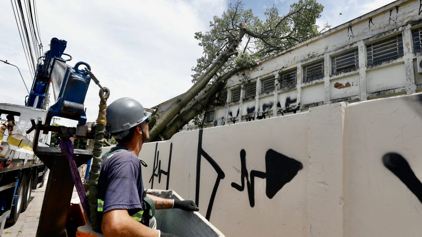Trabalhador com capacete em cesto de caminhão guindaste ao lado de árvore que caiu sobre o muro de um prédio em Porto Alegre, cidade no aviso laranja de tempestade de 28 de setembro