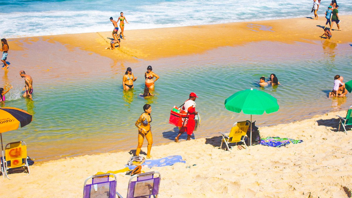 Banhistas na areia clara e na água rasa de uma praia do Rio de Janeiro, com um guarda-sol verde aberto e cadeiras de praia sob sol forte
