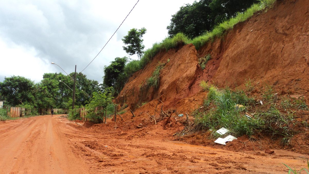 Barranco de terra vermelha que desabou sobre uma rua de terra, com cerca caída e lixo espalhado no pé da encosta