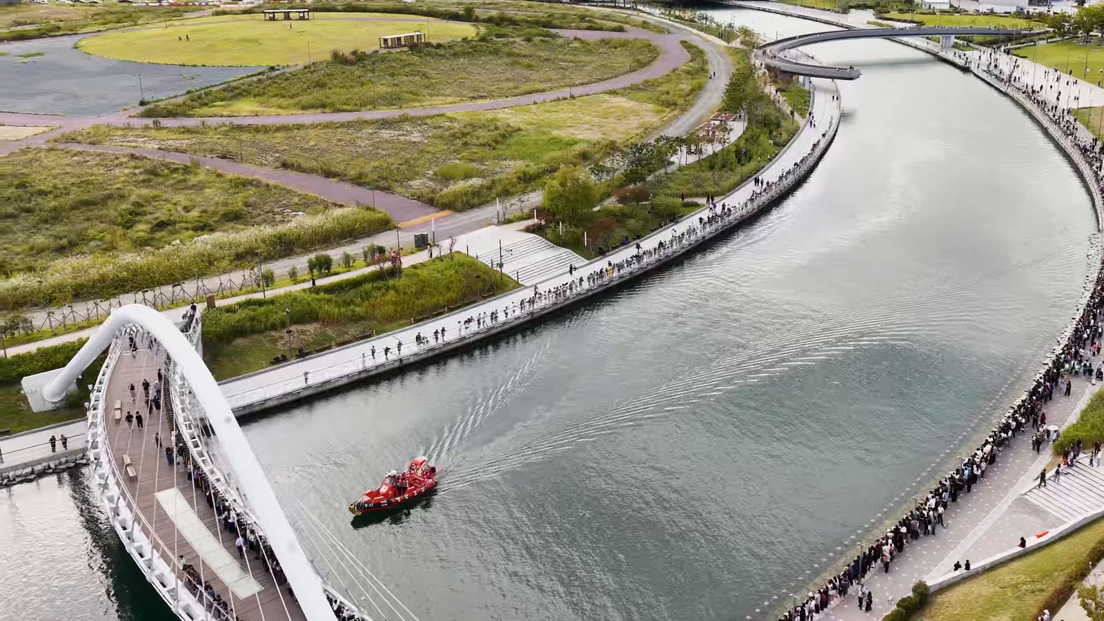 Canal em curva no parque do Porto Norte de Busan, com ponte em arco branca, um barco vermelho na água e as margens tomadas de gente