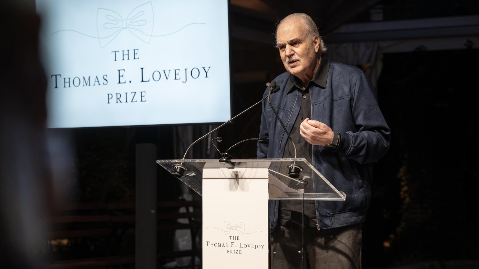 Carlos Nobre, de cabelo branco e jaqueta azul, discursa num púlpito de acrílico diante de um telão com o nome do Prêmio Thomas E. Lovejoy