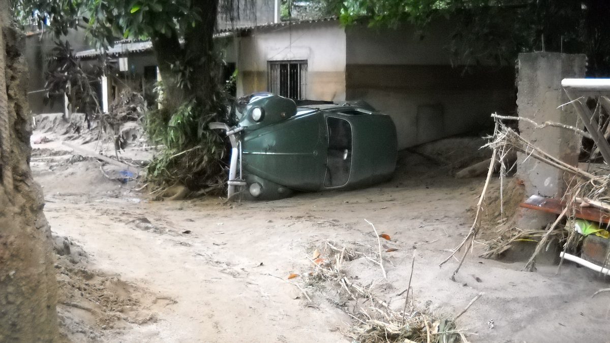 Carro verde tombado de lado em rua de terra coberta de lama e galhos, com casas ao fundo, depois de uma enxurrada em Xerém