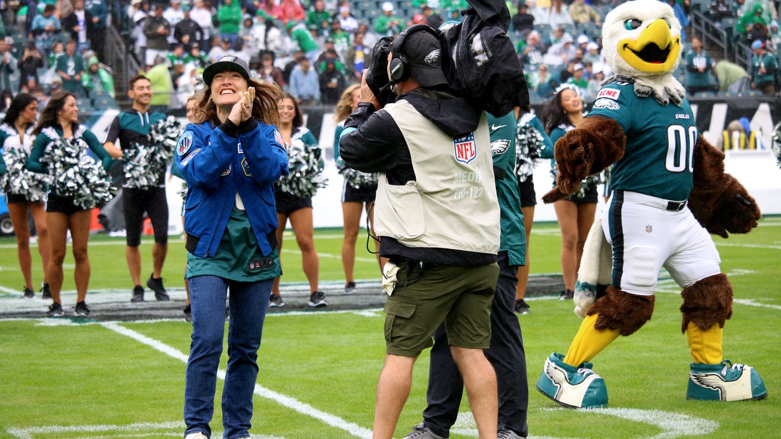 Christina Koch, astronauta da Artemis II, aplaude de casaco azul no gramado do estádio dos Eagles, ao lado de cheerleaders e do mascote