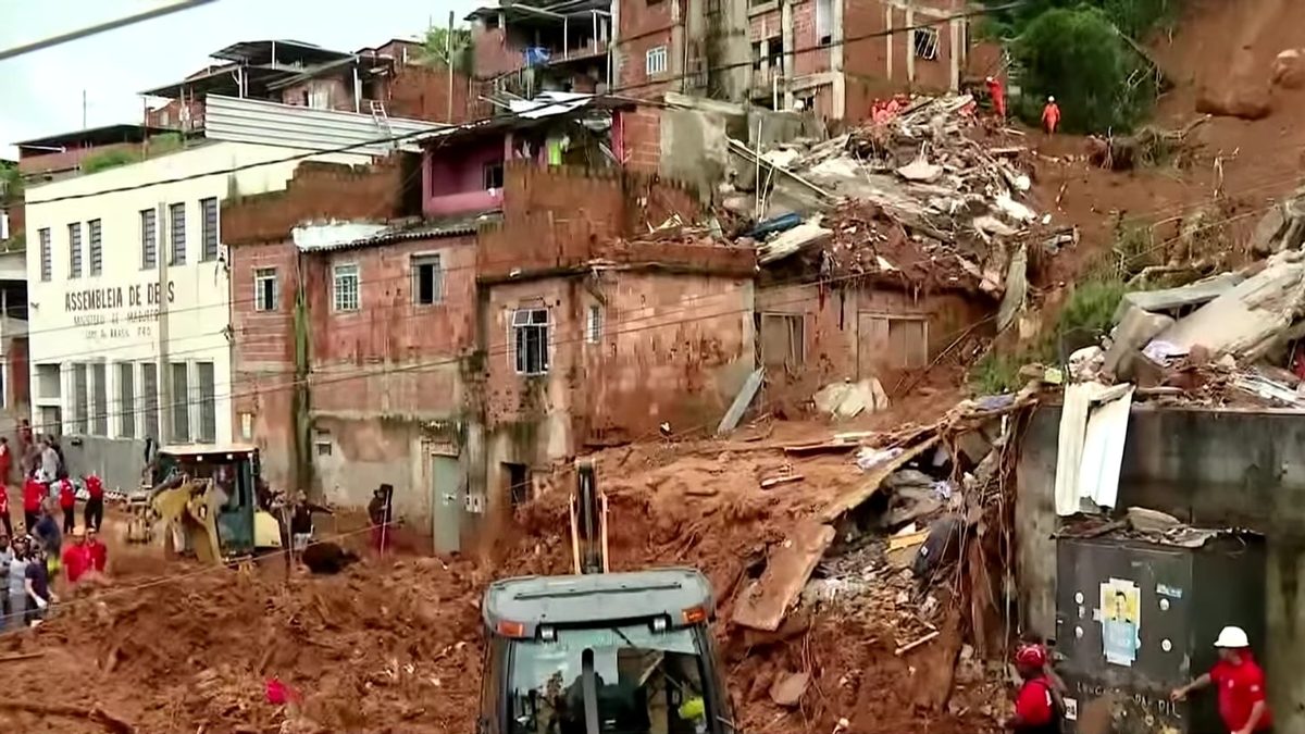 Terra vermelha e entulho cobrem uma rua em ladeira, com equipes de resgate e moradores diante das casas atingidas por deslizamento