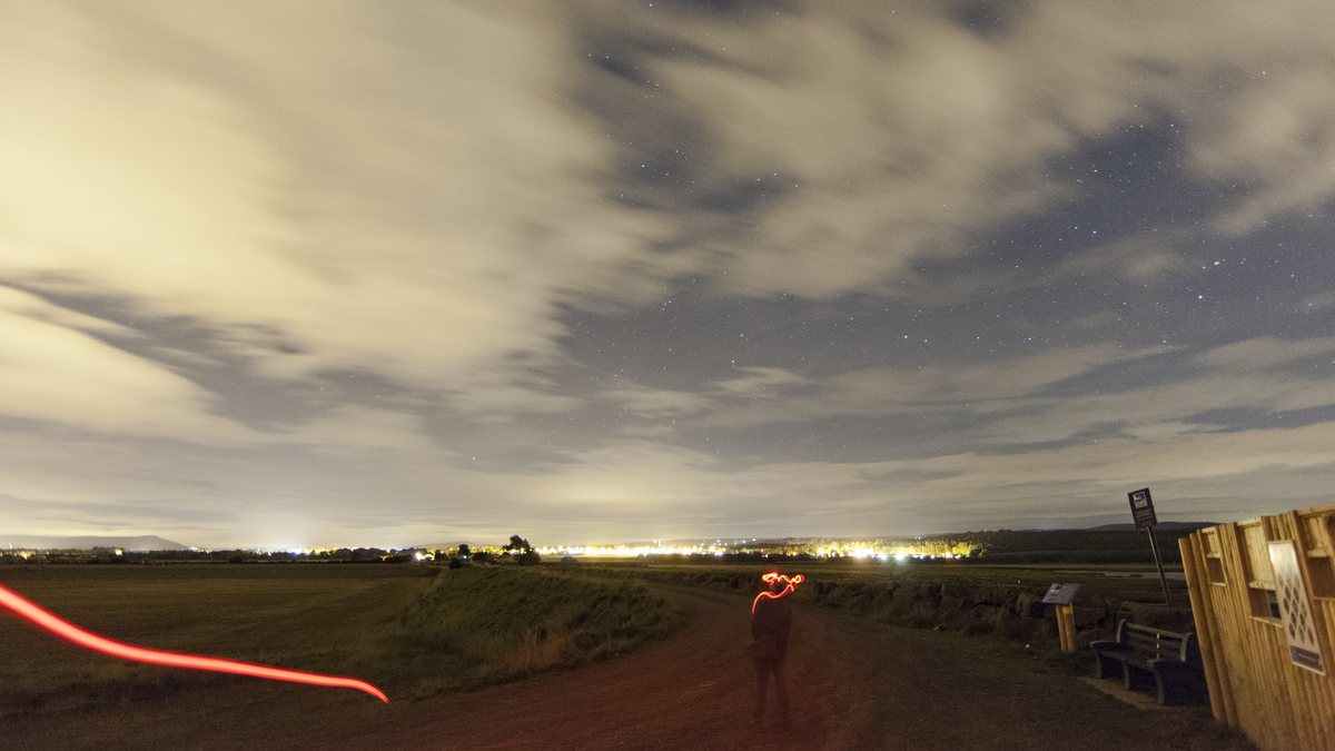 Estrada de terra sob céu coberto de nuvens à noite, com luzes de cidade no horizonte e uma pessoa deixando um rastro de luz vermelha, na noite das Dracônidas de 2015
