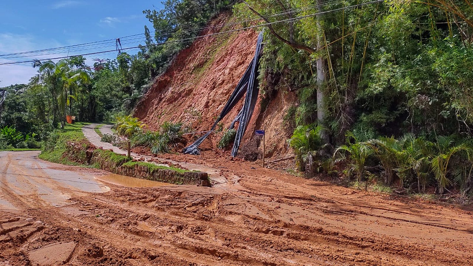 Encosta de terra vermelha que cedeu sobre a rua, com fios pendurados no barranco, um dos sinais de deslizamento e de risco depois de chuva forte