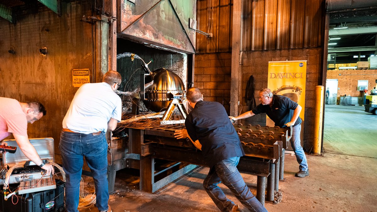 Quatro homens empurram a sonda DAVINCI sobre trilhos para dentro do forno, enquanto um deles opera um equipamento de medição