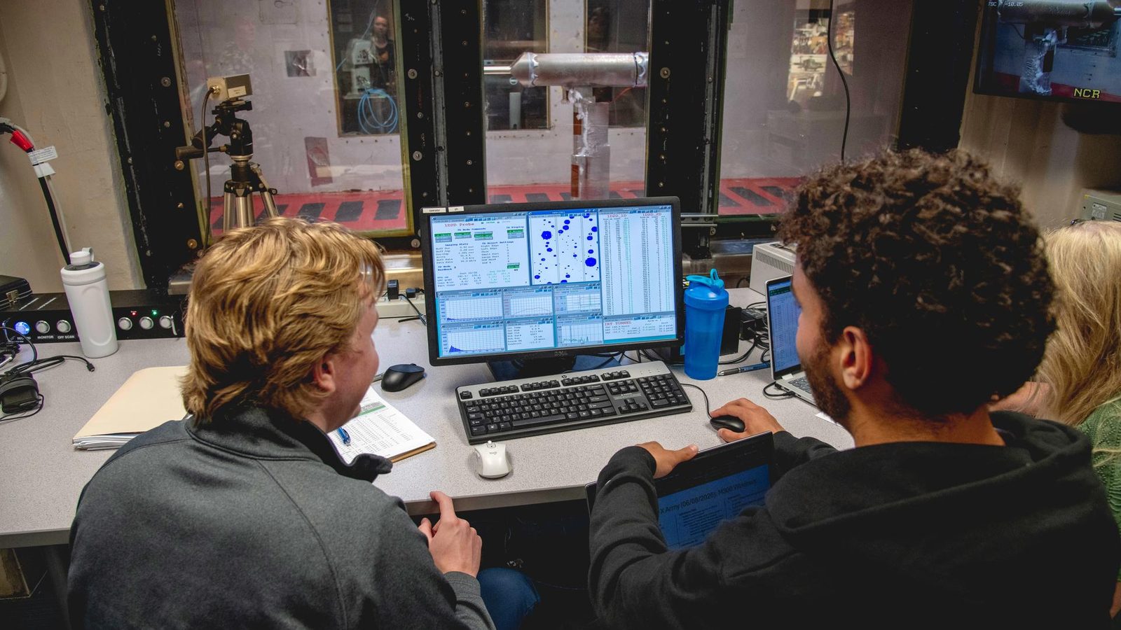 Dois engenheiros diante de um monitor com o tamanho das gotas do túnel, e a sonda coberta de gelo vista pela janela ao fundo