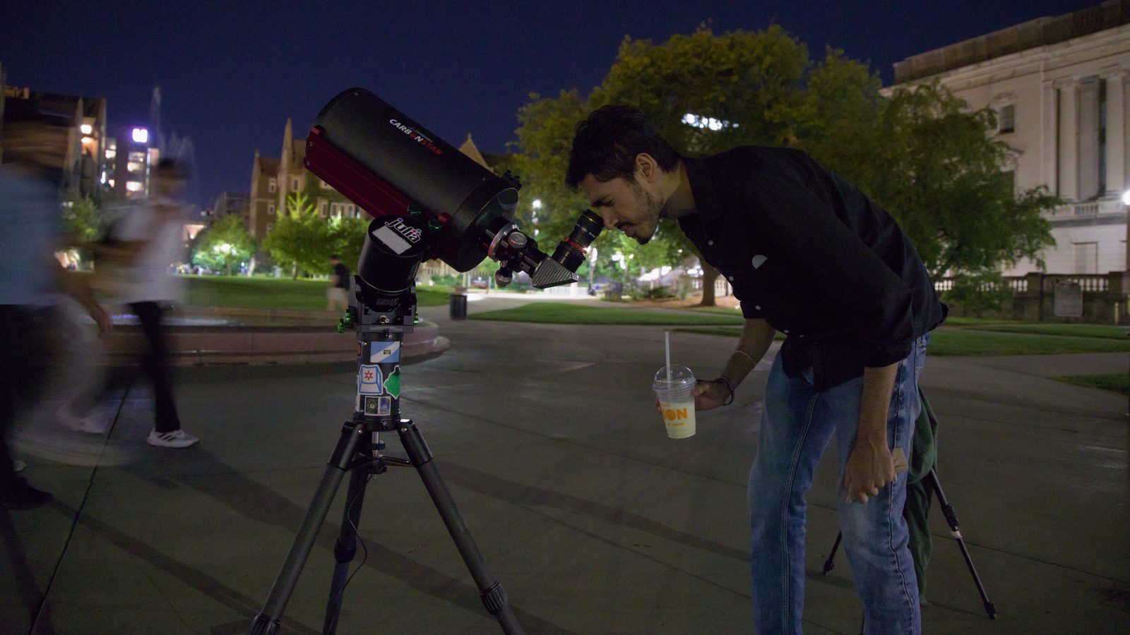 Estudante olha por telescópio de 8 polegadas numa praça à noite para ver Saturno, enquanto os anéis de Saturno com binóculo não aparecem separados