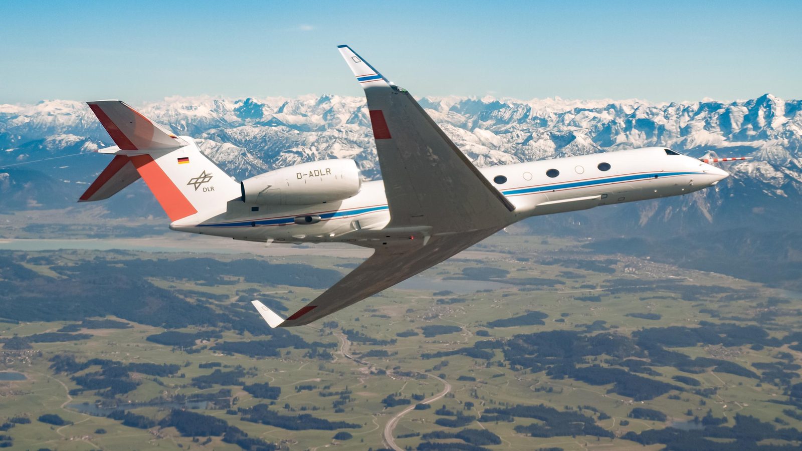 O avião-laboratório alemão HALO, um jato branco do DLR com listra azul, em voo sobre montanhas e campos, com a haste de medição no nariz