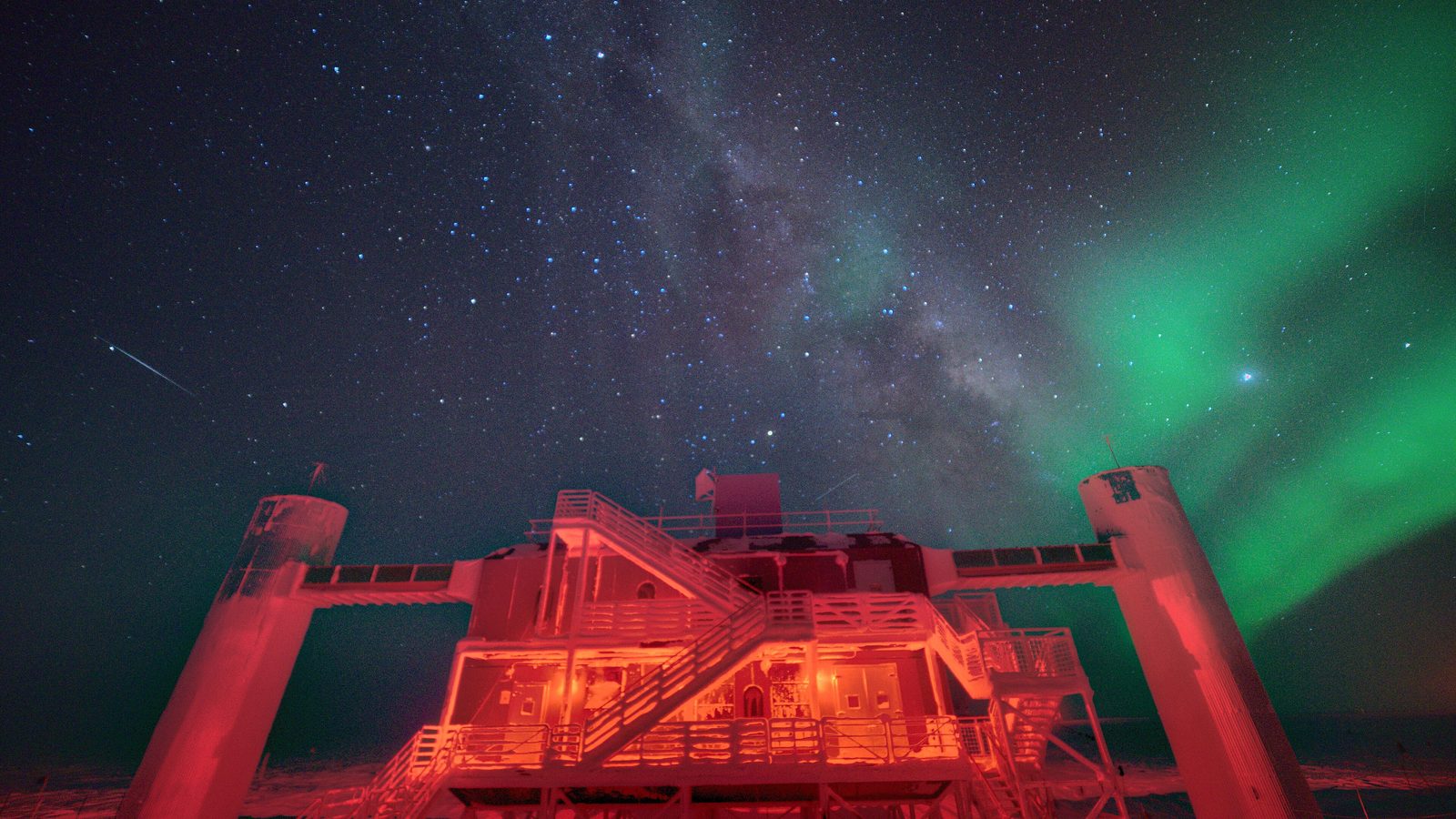 Laboratório do IceCube no Polo Sul à noite, iluminado de vermelho, sob a Via Láctea e uma aurora verde, no ano do Nobel de Física 2026