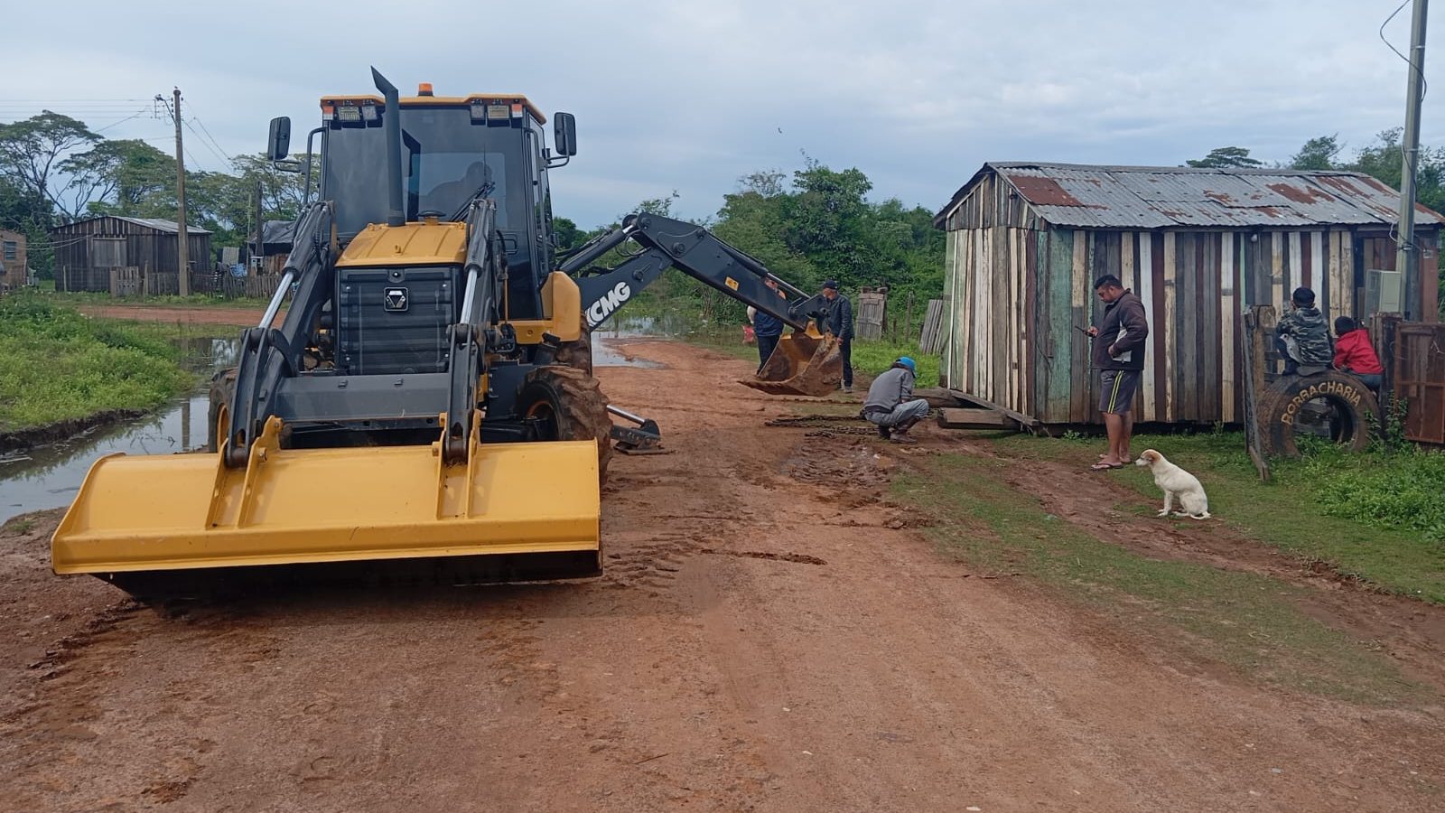 Retroescavadeira amarela e moradores numa rua de terra com água ao lado em Itaqui, no Rio Grande do Sul, na cheia do rio Uruguai