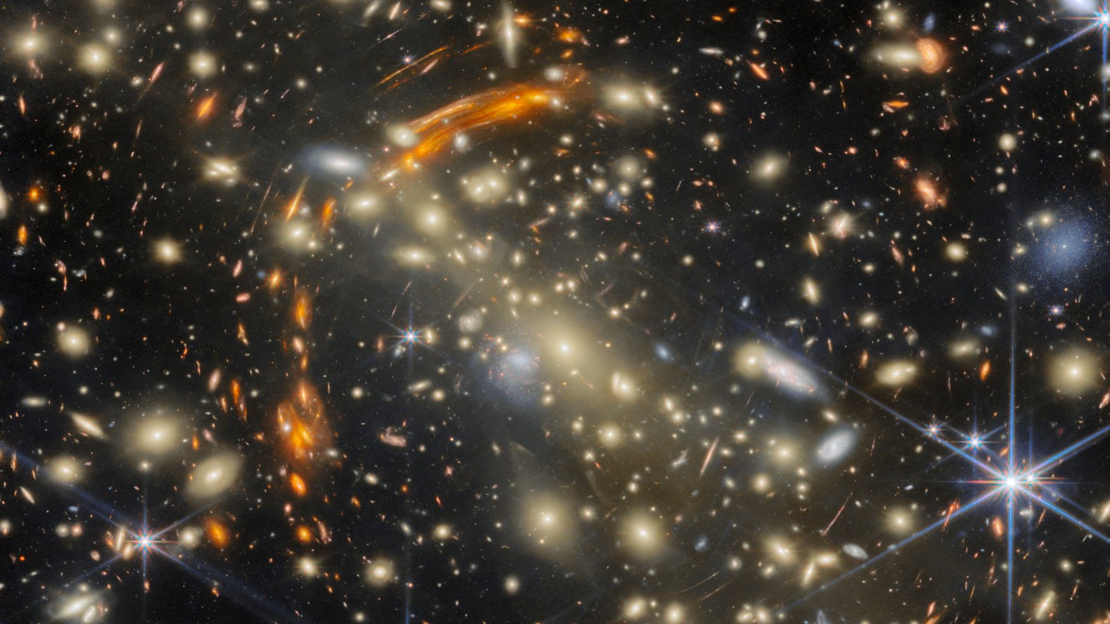 Imagem do James Webb com lente gravitacional: aglomerado de galáxias douradas e arcos alaranjados de galáxias distantes esticadas pela gravidade, com estrelas brilhantes de pontas azuis