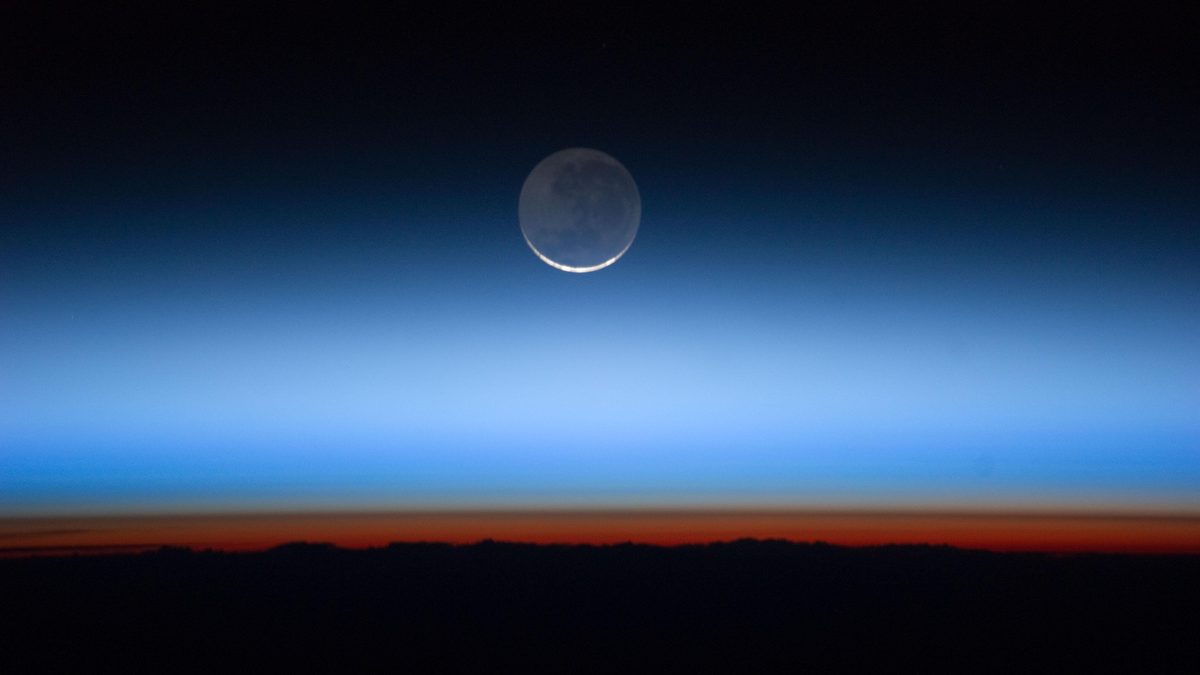 Borda da Terra vista da órbita, com faixa laranja-avermelhada junto à superfície, camadas de azul claro acima, o preto do espaço e uma Lua crescente no alto